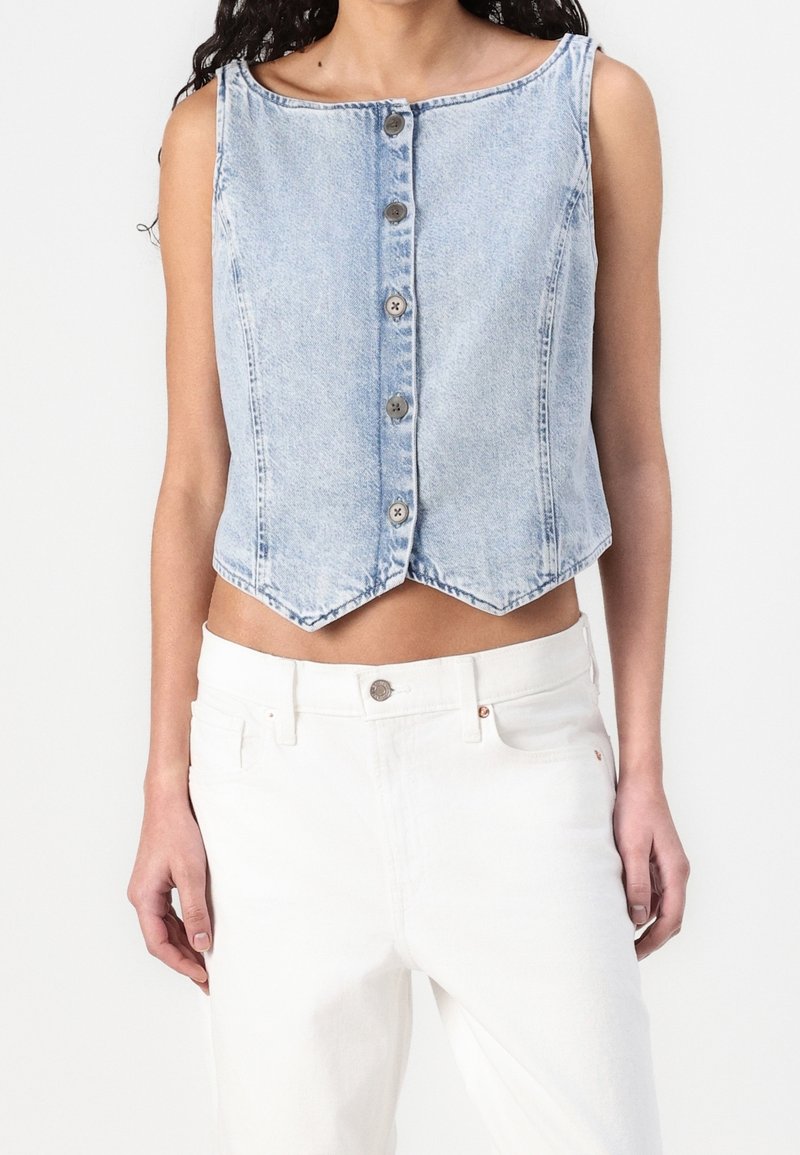 Veste en denim bleu clair sans manches avec cinq boutons devant, associée à un pantalon blanc taille haute porté par une personne.
