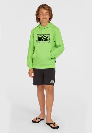 Hoodie vert fluo avec un logo graphique noir, associé à un short noir avec un logo blanc. Le modèle porte des tongs.