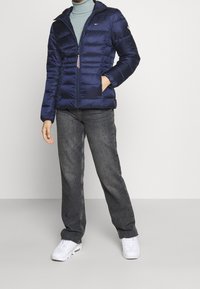 Giacca imbottita blu navy con zip frontale, cappuccio e collo a costine. Presenta cuciture orizzontali e due tasche frontali, abbinata a jeans grigi.