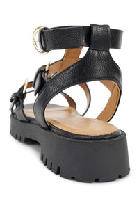 Schwarze Ledersandale mit einer dicken, strukturierten Plattformsohle, verstellbarem Knöchelriemen und dekorativen Schleifenakzenten. Sichtbare Beschläge in Goldtone.