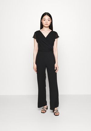 Combinaison noire avec des manches courtes agrémentées de dentelle, design enveloppant et coupe patte d'eph. Texture lisse, détails minimalistes, associée à des sandales noires à lanières.