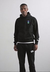 Felpa nera con cappuccio e tasca frontale, caratterizzata da un piccolo logo colorato. Abbinate a pantaloni sportivi neri con testo bianco.