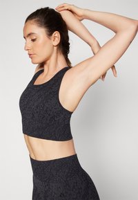 Crni sportski crop top s teksturiranim uzorkom leoparda, s okruglim izrezom i širokim naramenicama, u paru s odgovarajućim visokim tajicama.