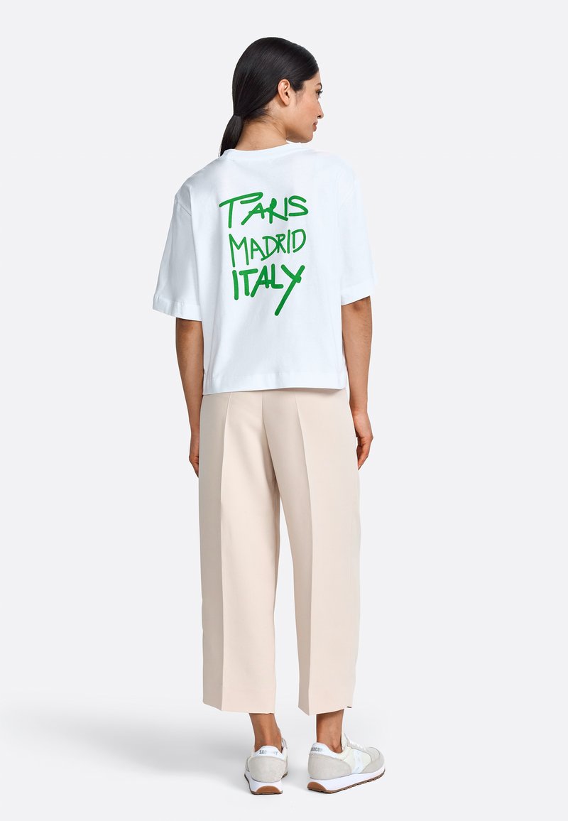 White cotton t-shirt with green text "PARIS MADRID ITALY" on the back. Loose fit, short sleeves, paired with beige pants and white shoes.