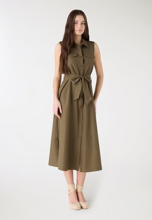 Giovane donna con lunghi capelli castani che indossa un vestito midi verde oliva senza maniche con bottoni frontali e vita annodata, abbinato a scarpe beige con lacci alla caviglia.