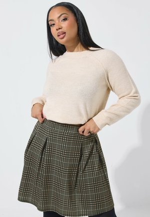 Vrouw met lang zwart haar, gekleed in een beige trui en geplooide donkergroene en bruine geruite rok, staand tegen een witte achtergrond.