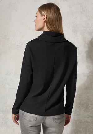 Schwarzes Langarmoberteil mit hohem Kragen, weißen Seitenstreifen, weichem Material und figurbetontem Schnitt, kombiniert mit grauen Jeans.