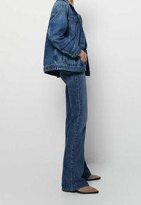 Denim jacket dengan nuansa biru, potongan santai, dan saku depan besar, dipadukan dengan celana jeans flared biru dan sepatu datar runcing berwarna krem.