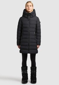 Mustast polsterdatud mantel kõrge kraega ja tõmblukuga, küljetaskute ja aktsentõmblustega, kantud mustade lumesaabastega.