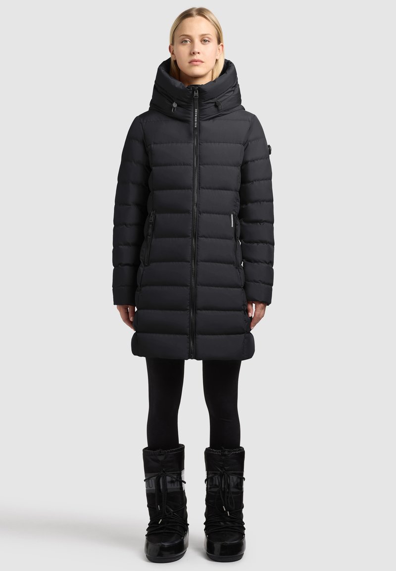 Mustast polsterdatud mantel kõrge kraega ja tõmblukuga, küljetaskute ja aktsentõmblustega, kantud mustade lumesaabastega.