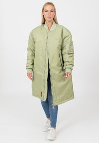 Lichtgroene lange bomberjack met een rits, geribde manchetten, zijzakken en een zak op de linkermouw. Gedragen over blauwe jeans en witte sneakers.