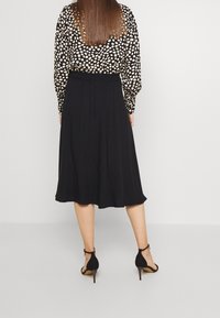 Schwarzer Rock mit fließendem Design, kombiniert mit einer Bluse mit langen Ärmeln, die ein cremefarbenes und schwarzes Punktmuster aufweist. Das Model trägt schwarze Sandalen mit Absatz.
