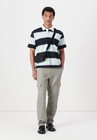 Pruhovaná polo košeľa v modrej a mätovej farbe, doplnená svetlosivými cargo nohavicami. Muž nesie čiernu crossbody tašku a má na sebe čierne topánky.