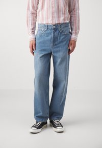 Homme portant un jean droit bleu clair, des baskets noires et blanches, et une chemise rayée rose et blanche à manches longues, se tenant contre un fond uni.