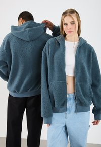 Blauwe textuur fleece hoodie met rits aan de voorkant, voorzien van een kap en twee voorzakken, gedragen met lichtblauwe spijkerbroek. Zwarte broek op de achtergrond.