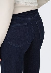 Gros plan d'une personne portant un jean denim bleu foncé taille haute avec poches arrière et un haut crème avec bordure en dentelle.