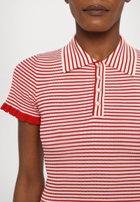 Czerwono-biała polówka w paski z żeberkową fakturą, krótkimi falbaniastymi rękawami i zapięciem na perłowy guzik. Kołnierzyk.