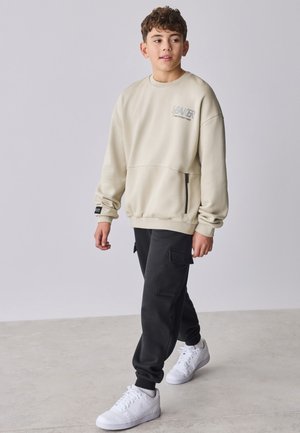 Garçon marchant à l’intérieur portant un sweat-shirt beige, un pantalon cargo noir et des baskets blanches, regardant légèrement vers sa gauche.