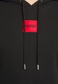 Felpa nera con cappuccio in tessuto morbido, caratterizzata da un'etichetta rettangolare rossa con la scritta "HUGO" in nero e lacci piatti neri con punte in argento.