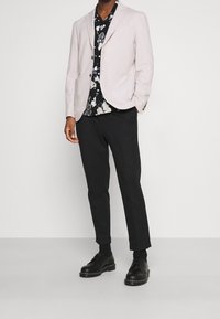 Blazer rosa claro sobre una camisa negra con flores, combinado con pantalones negros y zapatos de cordones negros. Tejido suave, ajuste entallado y líneas limpias.