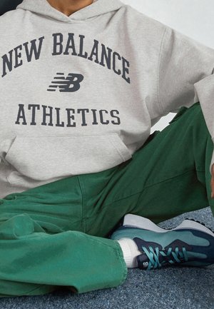 Henkilö istuu risti-istunnassa harmaa New Balance Athletics -huppari päällä, vihreät housut, valkoiset sukat ja sinivihreät New Balance -lenkkarit jalassa.