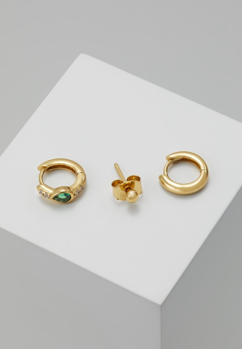 Deux boucles d'oreilles anneaux en or, l'une avec une pierre précieuse verte en forme de goutte et de petites pierres claires, et une boucle d'oreille simple en or avec un fermoir papillon sur une surface blanche.