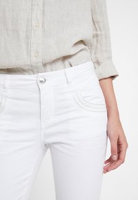 Mujer vestida con una camisa de lino de manga larga beige metida en unos jeans blancos ajustados con bolsillos frontales bordados, con la mano derecha relajada a un lado.