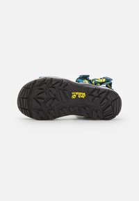 Svart gummisula med aggressivt slitmönster, med en gul logotyp. Ovandelen har blå och gröna remmar med justerbara stängningar.