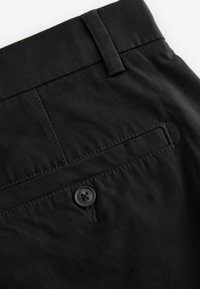 Primer plano de unos pantalones negros que muestran el bolsillo trasero con cierre de botón y un trabilla en la cinturilla.