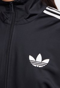 Chaqueta negra con cremallera hecha de tejido suave, con un cuello alto, tres rayas blancas en los hombros y un logo bordado en blanco.