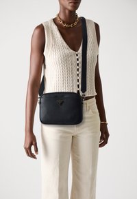 Borsa a tracolla nera in pelle con finitura texturizzata, hardware argentato e logo triangolare. Indossata con un gilet lavorato a maglia color crema e pantaloni beige.