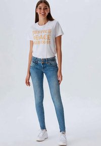White t-shirt with yellow text reading "Justice, Peace, Freedom" paired with blue skinny jeans and white sneakers. Casual outfit, simple design.