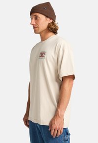 Beige short-sleeve T-shirt with front graphic design; relaxed fit. Worn with light blue denim pants and a brown knit beanie.