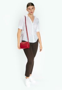 Camicetta bianca con maniche corte, pantaloni marroni aderenti e sneaker bianche. Tiene una piccola borsa a tracolla rossa con superficie testurizzata.