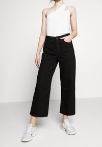 Schwarze weite Jeans mit hohen Taillenschnitt, kombiniert mit einem weißen Top mit einem Schulterträger. Das Model trägt weiße Sneaker und zeigt einen entspannten Look.