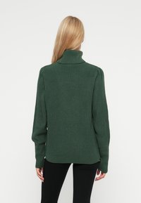 Suéter de malha verde-escuro com um gola alta, textura canelada, mangas longas e um corte relaxado. Detalhes de ombros franzidos são notáveis.