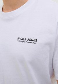 Valkoinen puuvillainen t-paita, jossa on musta brodeerattu logo rinnassa. Klassinen pyöreä kaula-aukko, lyhyet hihat ja sileä pinta.