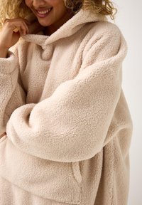 Przytulna, oversize'owa polarowa bluza z kapturem w jasnym beżowym kolorze. Miękka, teksturowana tkanina, z przednią kieszenią i luźnym fasonem. Ciepły i komfortowy design.