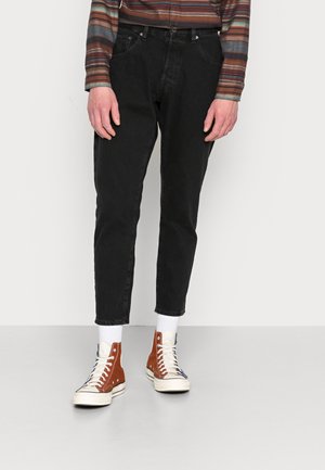 Person trägt schwarze, schmal zulaufende Jeans, braun gestreiftes langärmliges Hemd, weiße Socken und braune High-Top-Sneaker und steht vor einem schlichten Hintergrund.