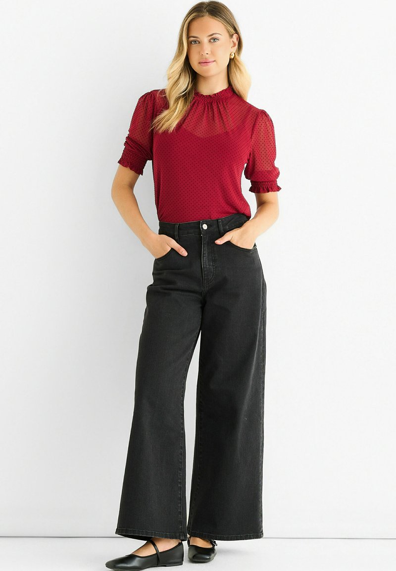 Red short-sleeve top with polka dots, high neck, and puffed sleeves paired with high-waisted, wide-leg black jeans. Black shoes.