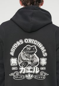 Sweat-shirt noir avec un graphisme d'une grenouille stylisée en veste, avec "ADIDAS ORIGINALS" et du texte japonais en blanc.