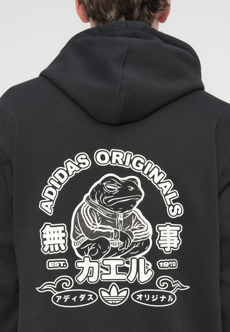 Schwarzer Pullover mit einer Grafik eines stilisierten Frosches in einer Jacke, mit "ADIDAS ORIGINALS" und japanischem Text in Weiß.