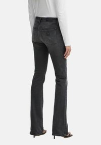 Jeans a zampa d'elefante in denim nero con finitura sbiadita. Presentano due tasche posteriori, un'etichetta con logo e un'altezza vita media. Indossati con una camicia bianca a maniche lunghe.
