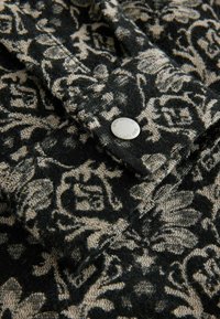 Schwarz-beige gemusteter Stoff mit floralem Design; mit einem silbernen Knopfdetail. Weiche Textur mit komplizierter Webart.