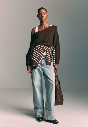 Brauner übergroßer Pullover, beige gestreifter Überwurf, hellblaue weit geschnittene Jeans, braune Ledertasche, dunkle Stiefel, minimale Accessoires.