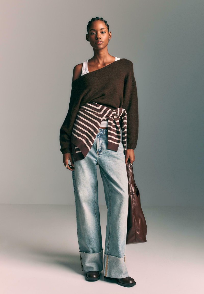 Bruine oversized trui, beige gestreepte wikkel, lichtblauwe wijde jeans, bruine leren tas, donkere laarzen, minimale accessoires.