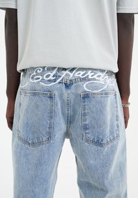 Světle modré denimové džíny s vyšitým logem "Ed Hardy" na pásku, tradičními zadními kapsami a uvolněným střihem.