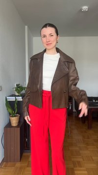 Νέα γυναίκα που στέκεται σε εσωτερικό χώρο, φοράει καφέ δερμάτινο μπουφάν, λευκό τοπ και φωτεινό κόκκινο παντελόνι με δεμένη ζώνη στη μέση, χαμογελώντας στην κάμερα.