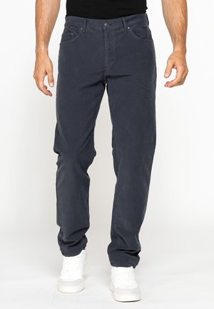 Uomo che indossa pantaloni dritti grigio scuro e sneakers bianche, in piedi davanti a uno sfondo bianco semplice.