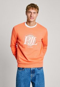 Sudadera naranja de cuello redondo, con un estampado gráfico blanco y el logo "Pepe Jeans". Combinada con unos jeans de mezclilla azul.